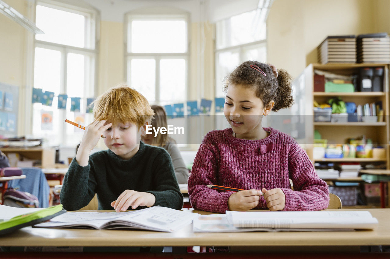 Smiling girl sitting by confused blond boy at desk in classroom