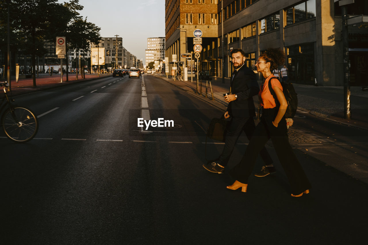 Female and male colleagues crossing street in city during sunset