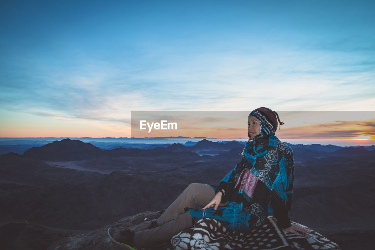 Woman looking away while sitting on mountain against sky during sunset