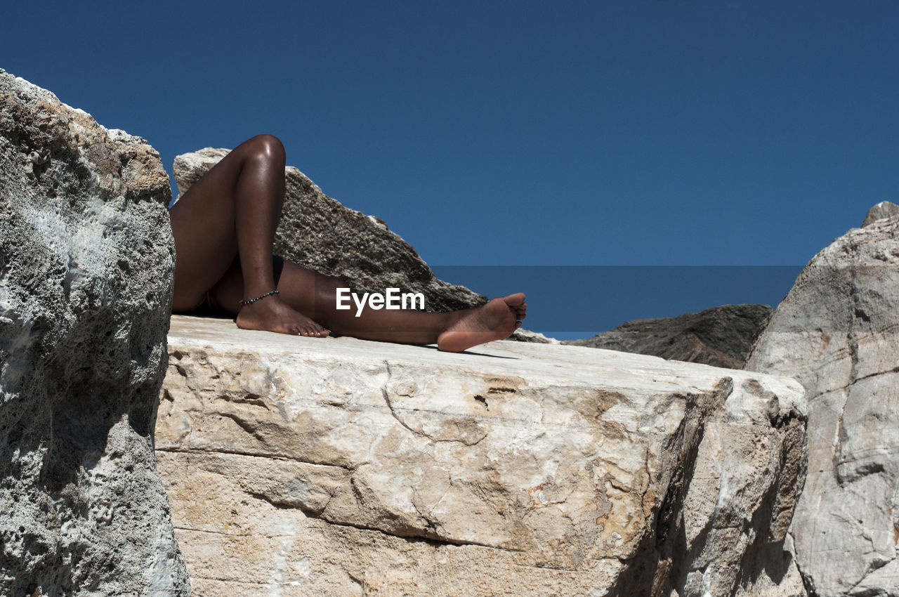 Low section of woman lying on rock against clear blue sky