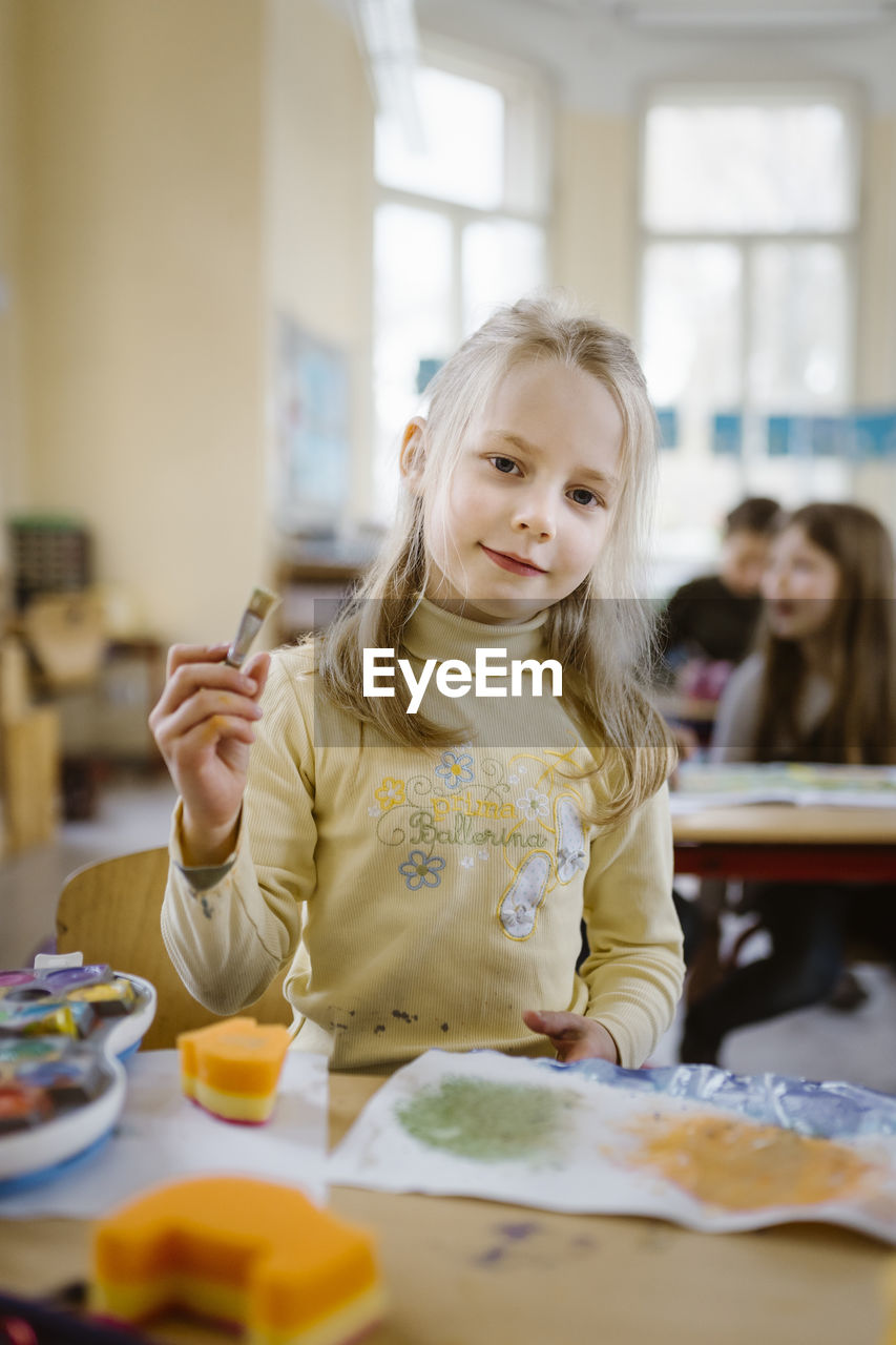 Portrait of female blond student holding paintbrush in classroom