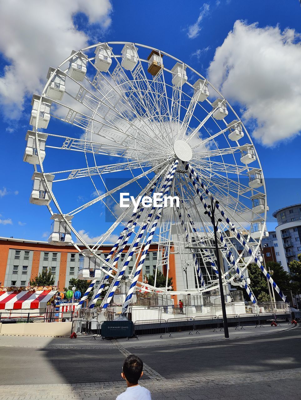 ferris wheel, amusement park ride, amusement park, arts culture and entertainment, architecture, sky, city, recreation, built structure, park, travel destinations, nature, cloud, leisure activity, carnival, traveling carnival, day, wheel, building exterior, outdoors, tourism, travel, amusement ride, landmark, person, transportation, circle, geometric shape, city life, tree, fun, blue, adult, shape, tourist