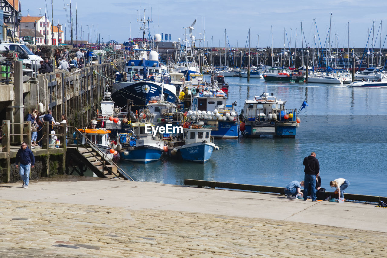 water, nautical vessel, transportation, sea, harbor, mode of transportation, coast, port, nature, architecture, pier, dock, sky, ship, infrastructure, moored, beach, group of people, boat, marina, sailboat, men, city, vehicle, body of water, land, travel, day, holiday, vacation, travel destinations, lifestyles, outdoors, trip, commercial dock, watercraft, adult, fishing, bay, tourism, crowd, built structure, mast, building exterior, women, pole, large group of people, sunlight, walkway