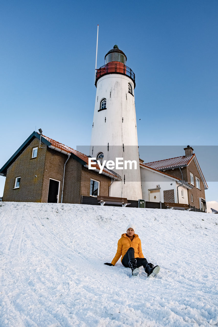 Full length of couple sitting on snow covered land against lighthouse