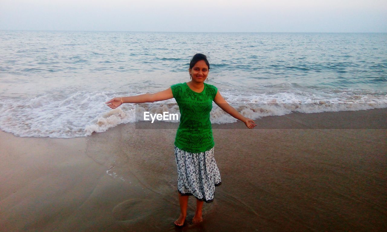 Portrait of smiling woman standing with arms outstretched on shore at beach against sky