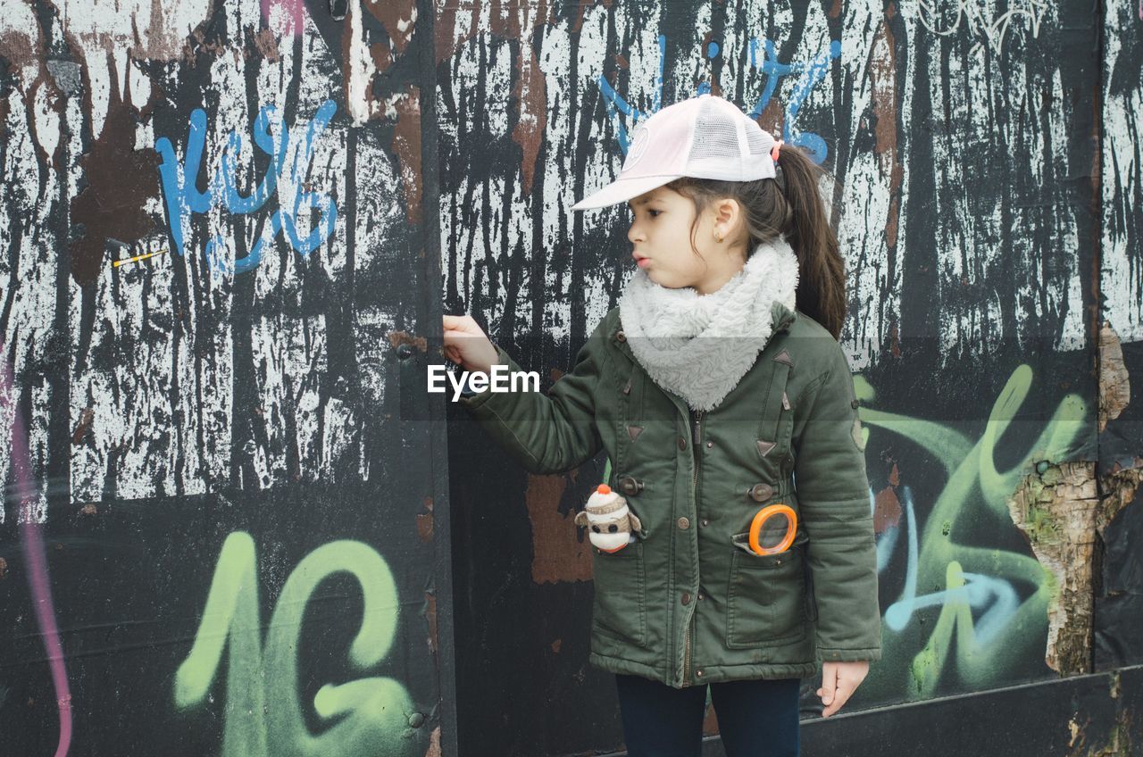 Girl wearing warm clothing and cap while standing against graffiti wall