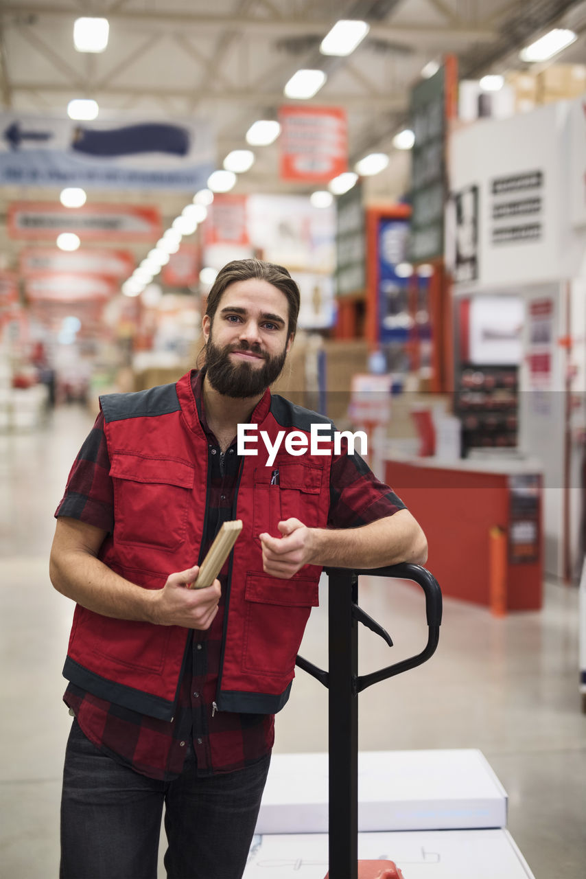 Portrait of confident salesman holding folding ruler while standing by handtruck in hardware store