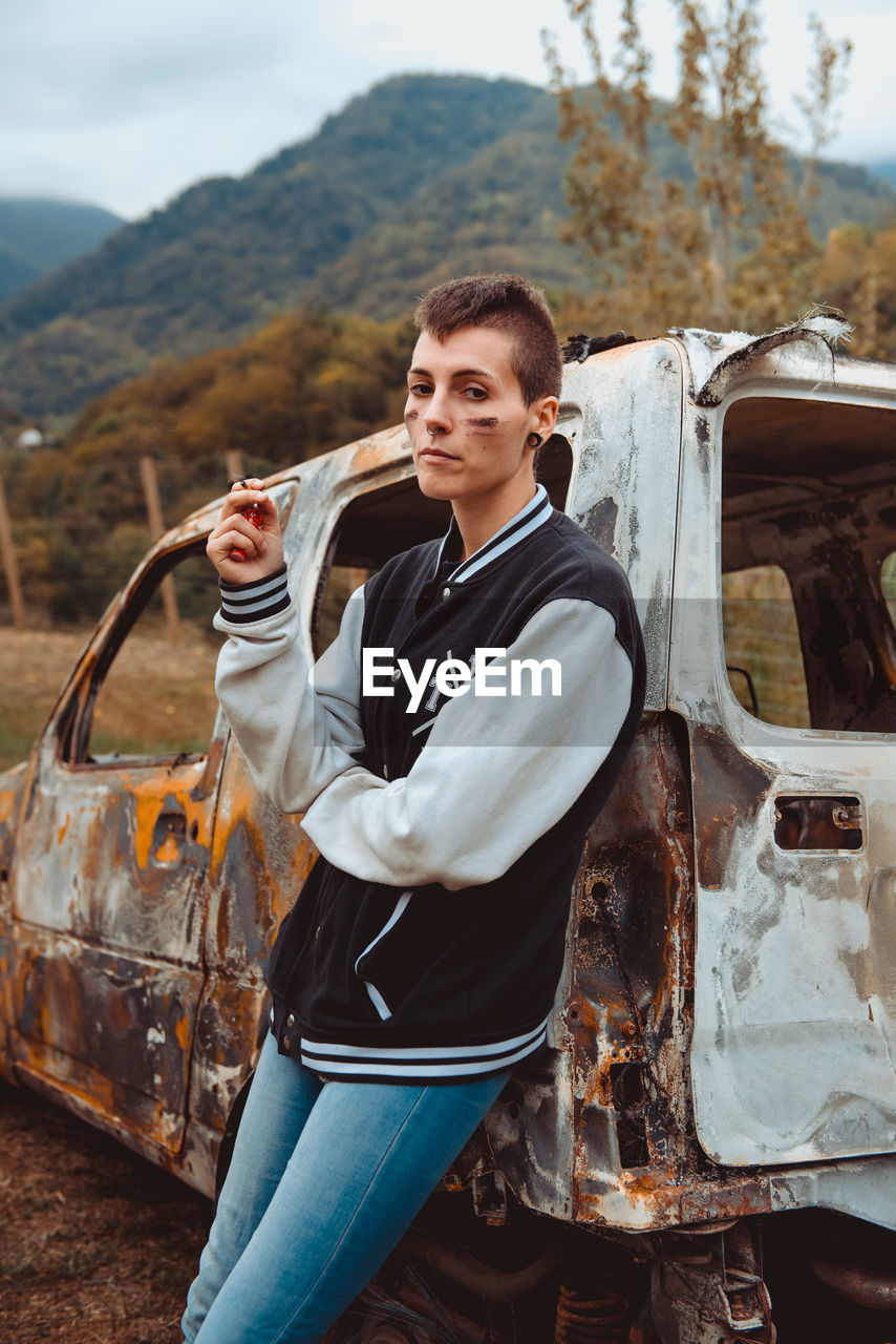 Young female with short hair smoking cigarette while resting near aged burnt vehicle in countryside looking at camera