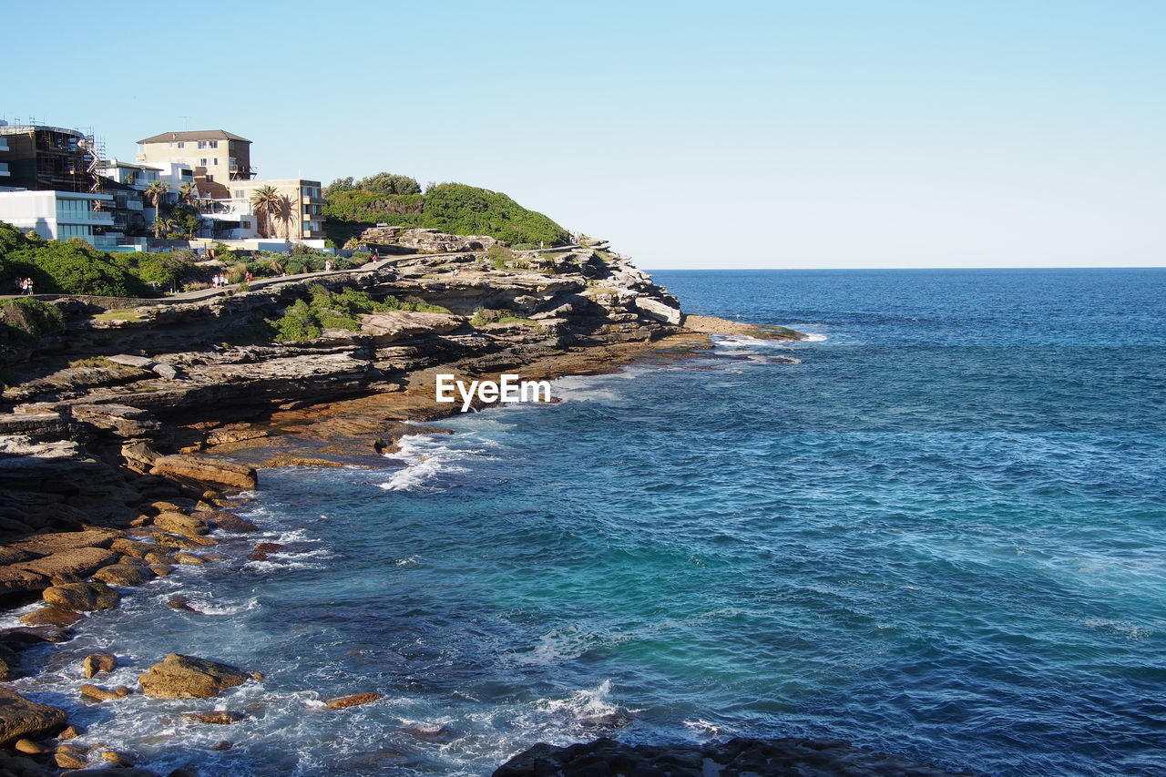 Buildings on rocky shore against clear blue sky