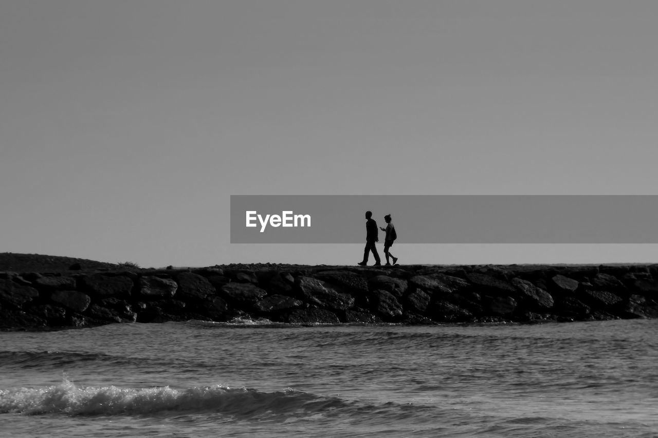 Silhouette friends walking on groyne by sea against clear sky