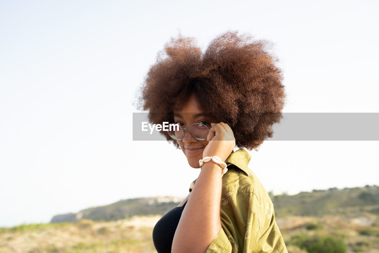 Side view of smiling young african american female with curly hair touching glasses and looking at camera while enjoying summer holidays in countryside