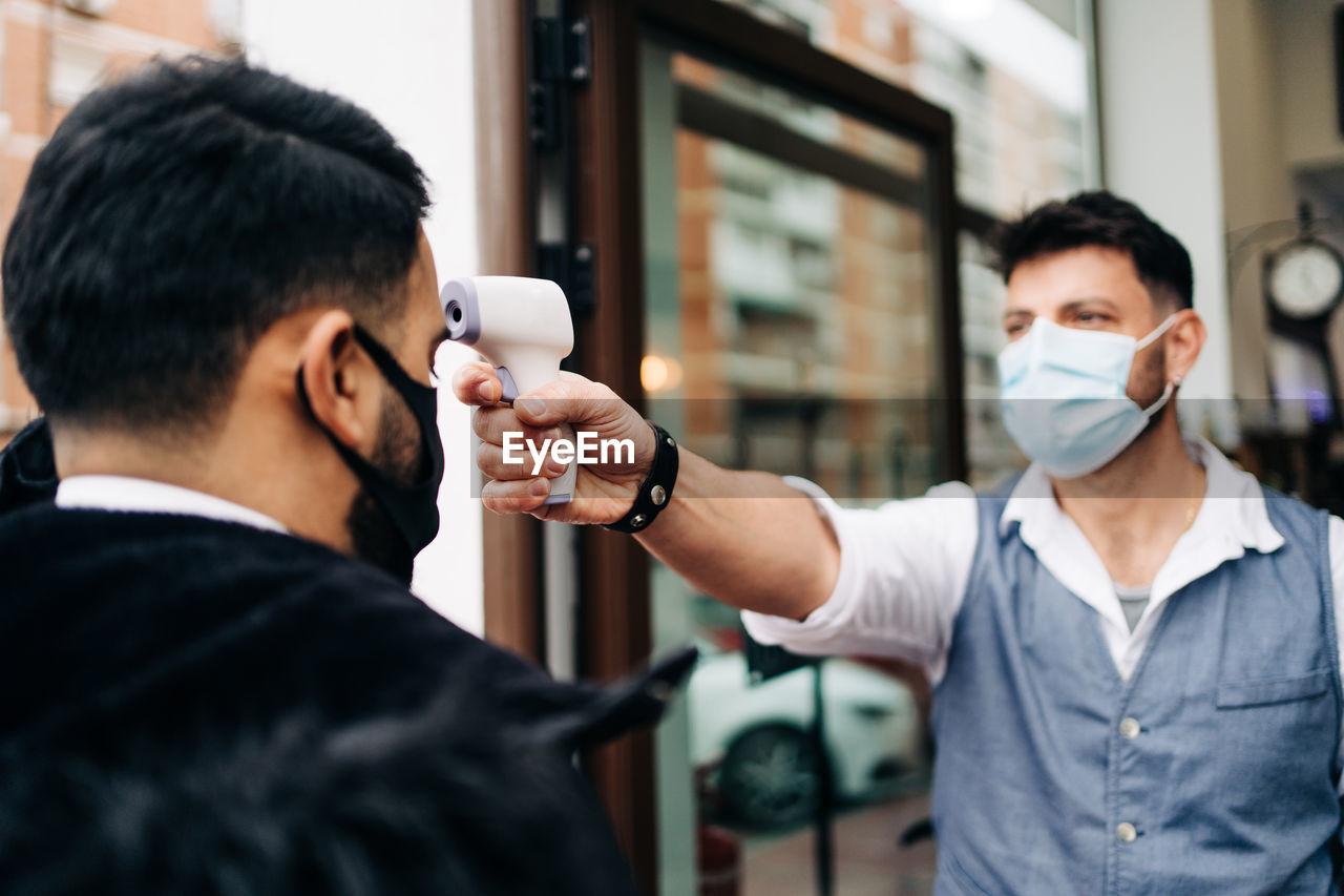 Male hairdresser in sterile mask measuring temperature of crop anonymous colleague with infrared thermometer at door of barbershop