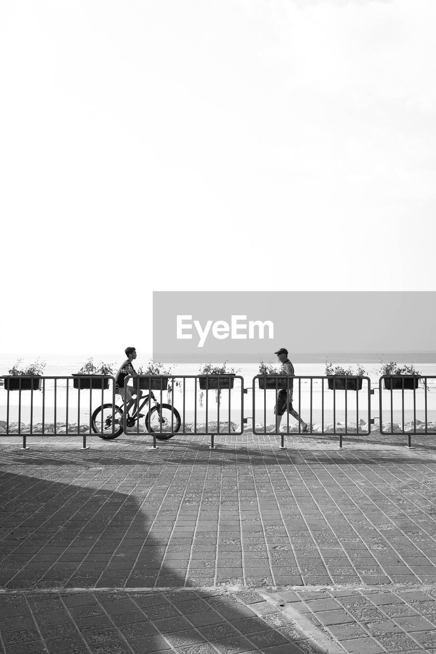 Man walking while teenager boy riding bicycle by railing against sea