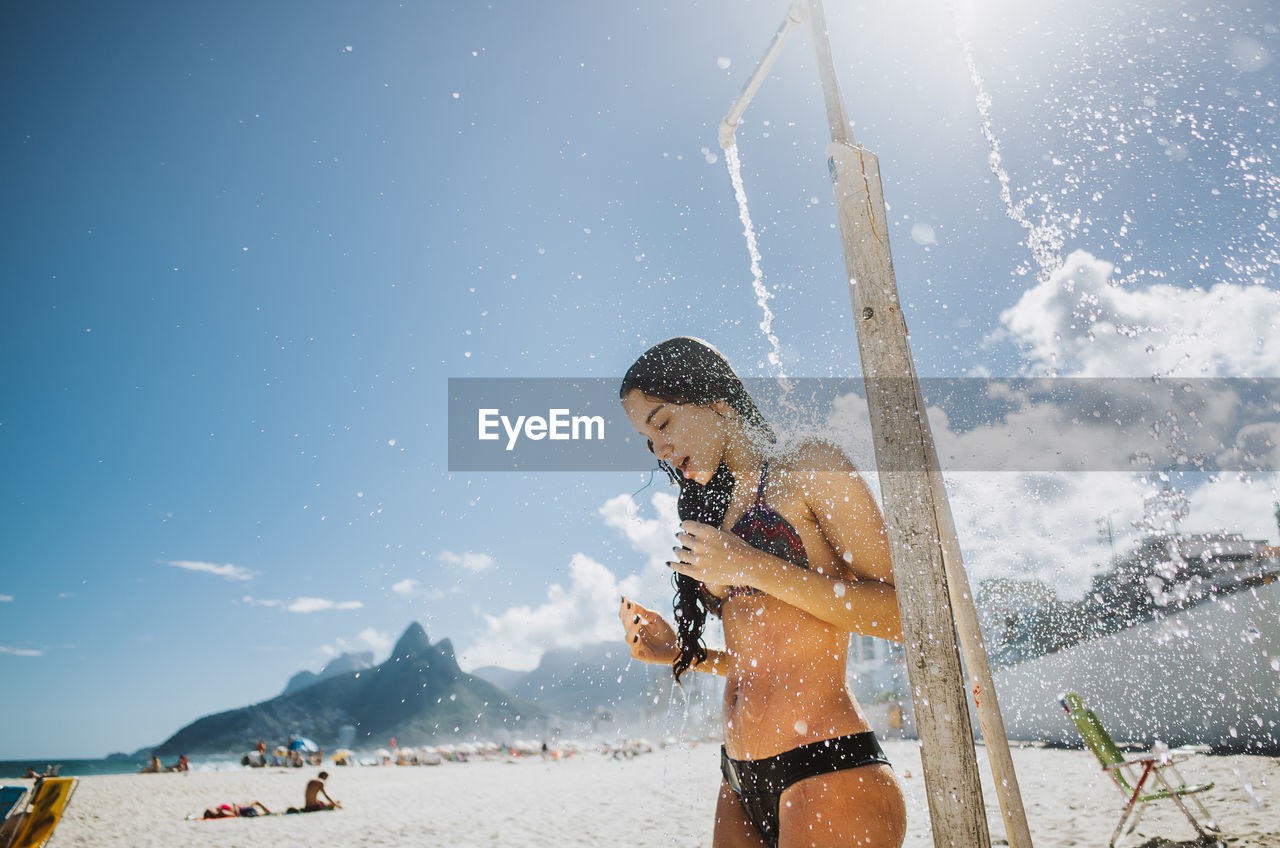 Woman bathing below shower at beach against sky