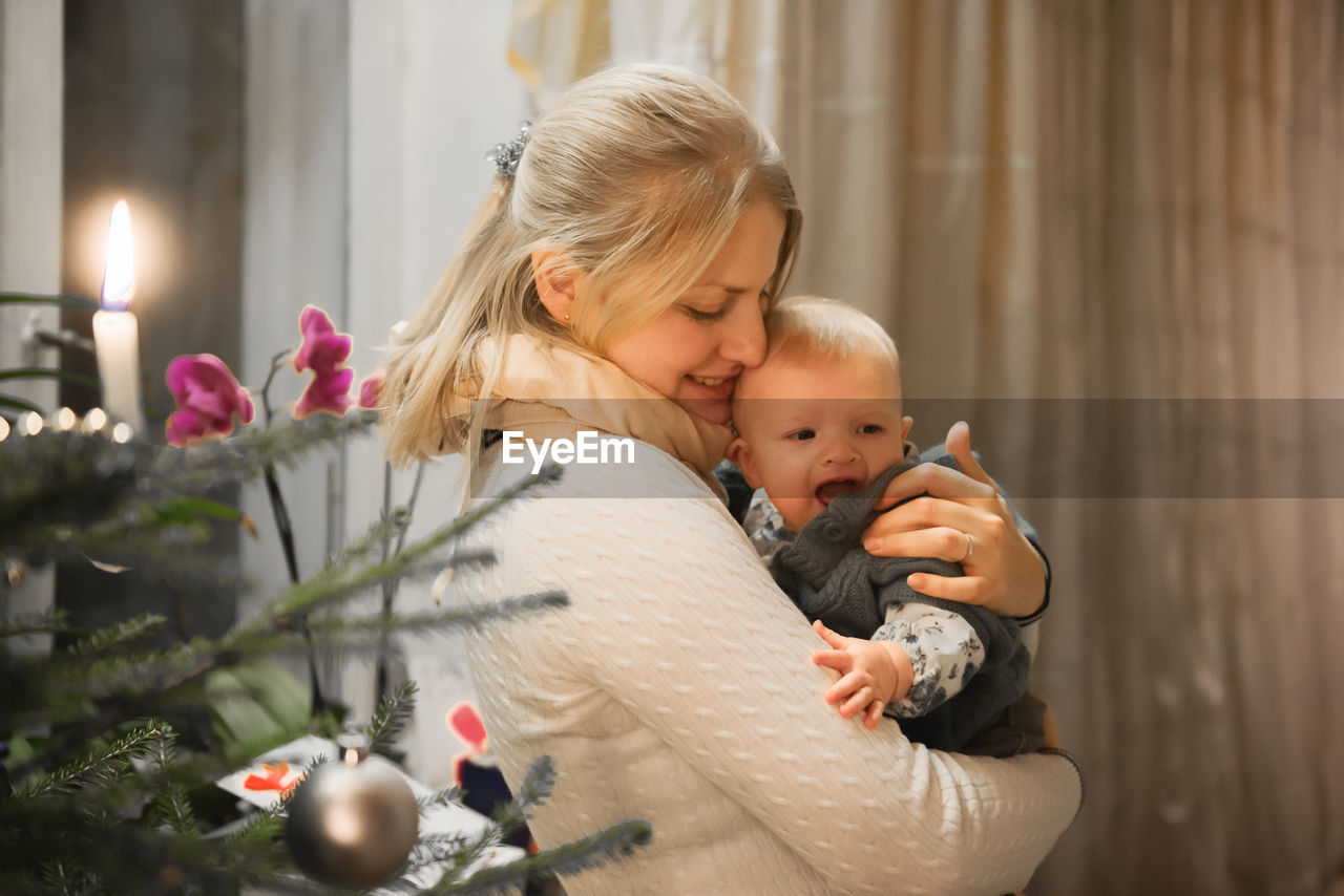 Side view of mother carrying baby girl by christmas tree at home