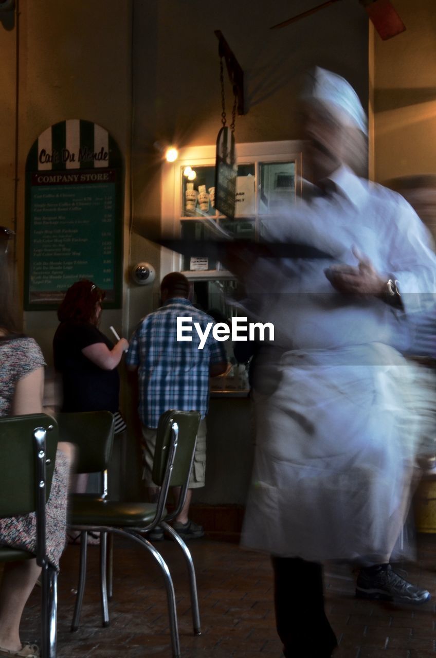 REAR VIEW OF MAN SITTING ON TABLE AT ILLUMINATED CAFE