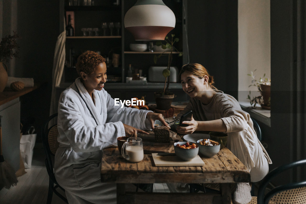 Smiling non-binary person sharing smart phone with woman having breakfast at home