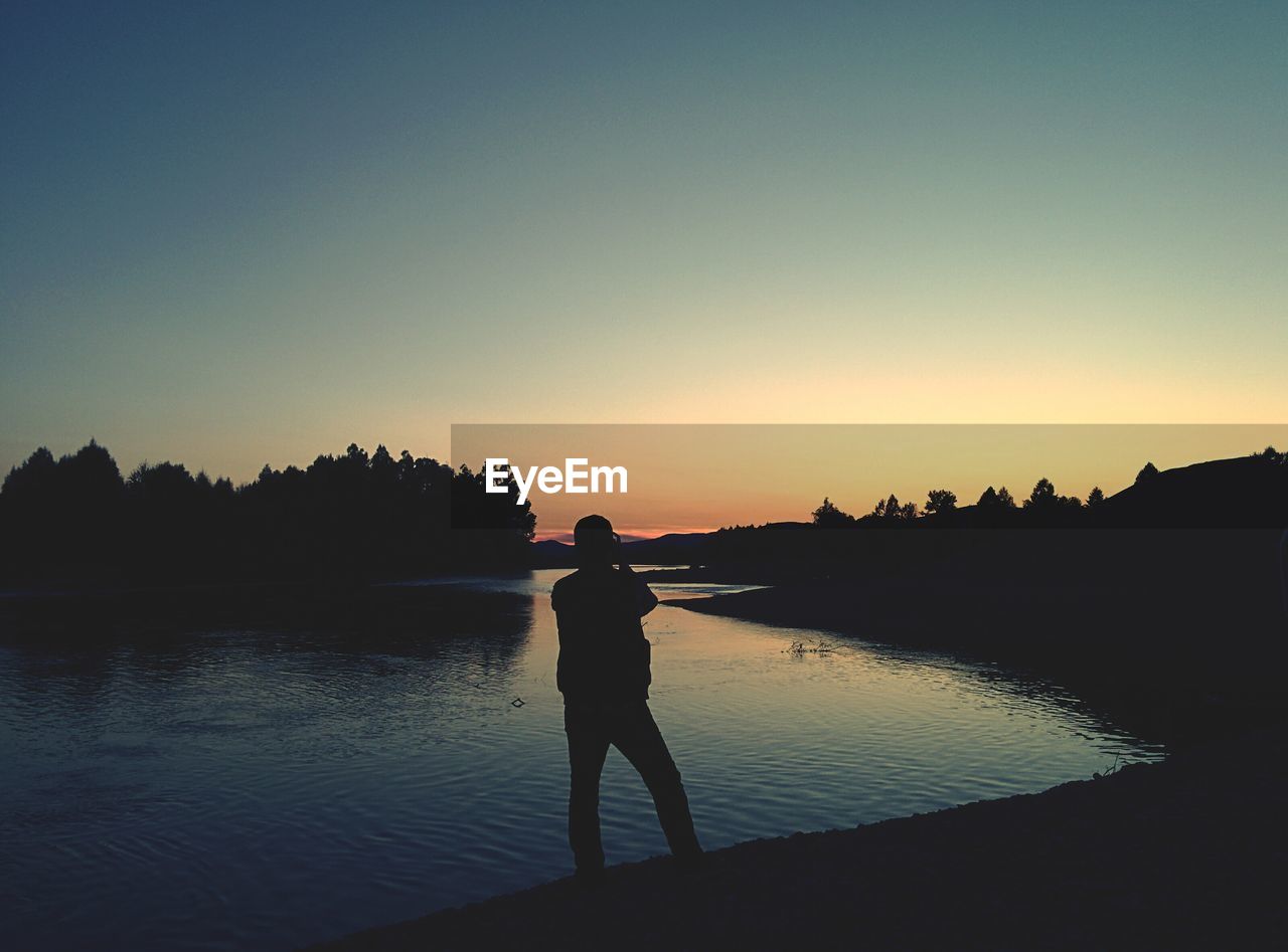Silhouette man standing by river against clear sky during sunset