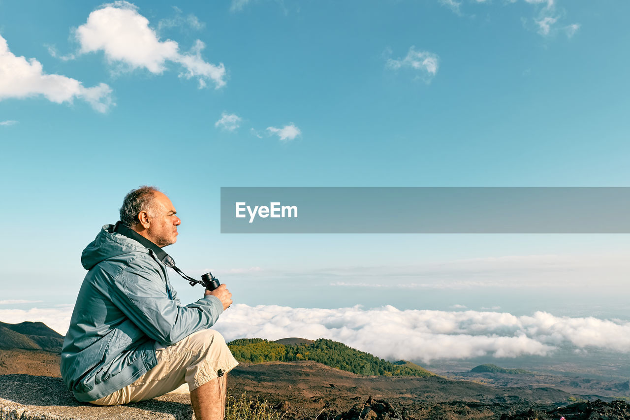 Mature bearded man looking through binoculars at panoramic view of summits of active volcano etna.