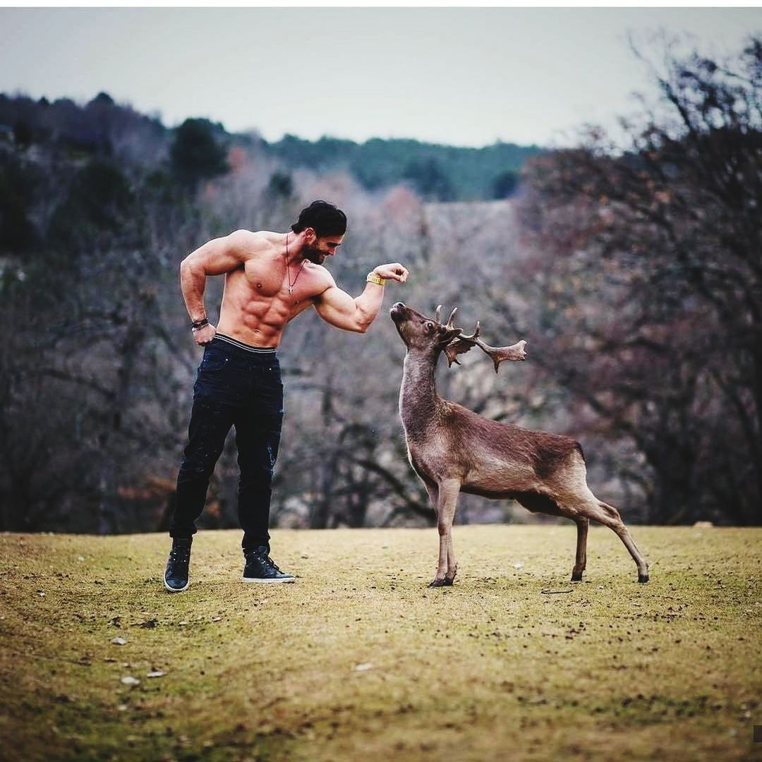 Shirtless man with muscular build standing by deer on field