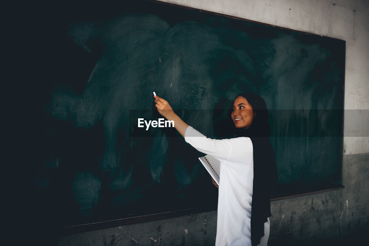 Side view of teacher writing on blackboard in classroom