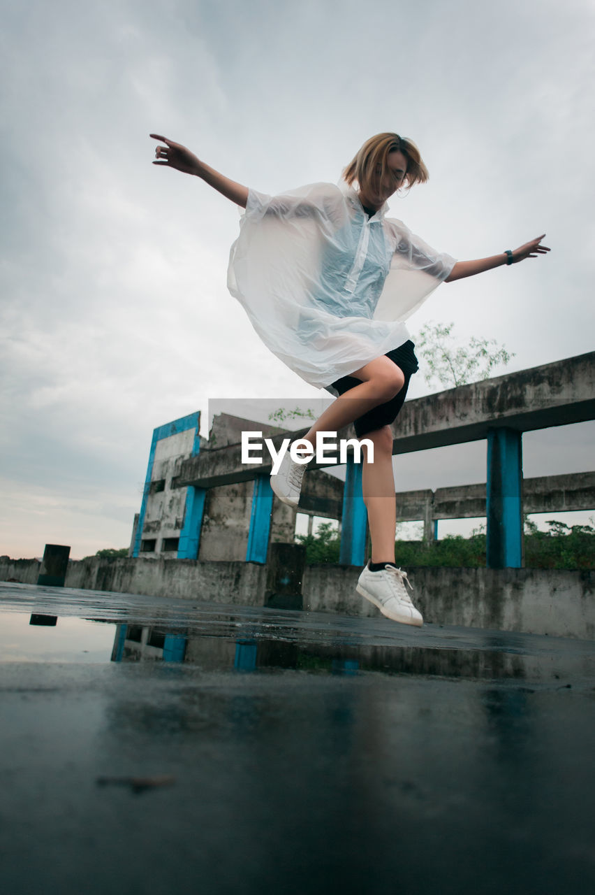 Full length of young woman wearing raincoat while jumping over puddle against sky