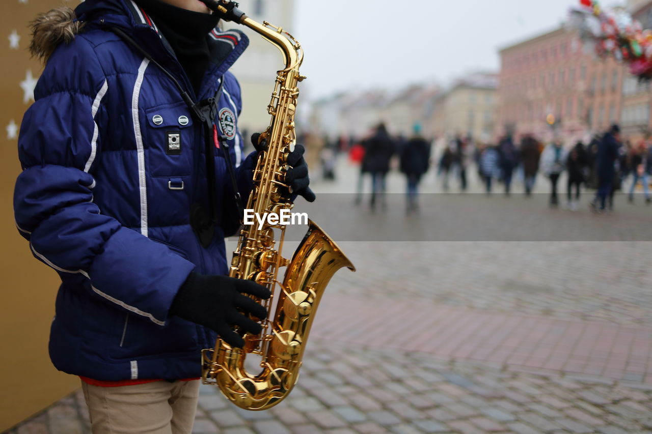 saxophone, musical instrument, music, arts culture and entertainment, musician, saxophonist, woodwind instrument, focus on foreground, performance, street, one person, city, holding, standing, adult, outdoors, street musician, wind instrument, skill, musical equipment, clothing, day, person, street artist, brass instrument, architecture, crowd, men