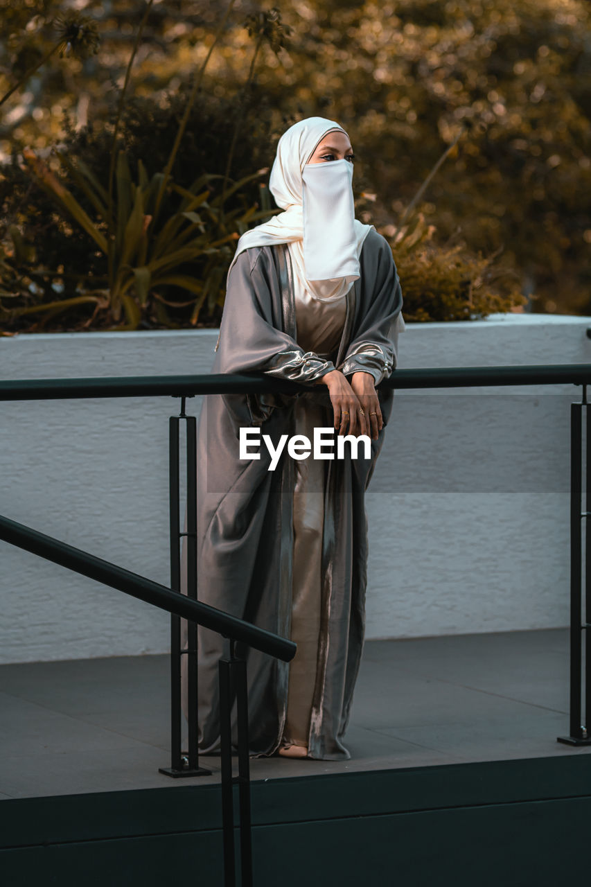 Full length picture of a veiled woman with white niqab and hijab standing by railing
