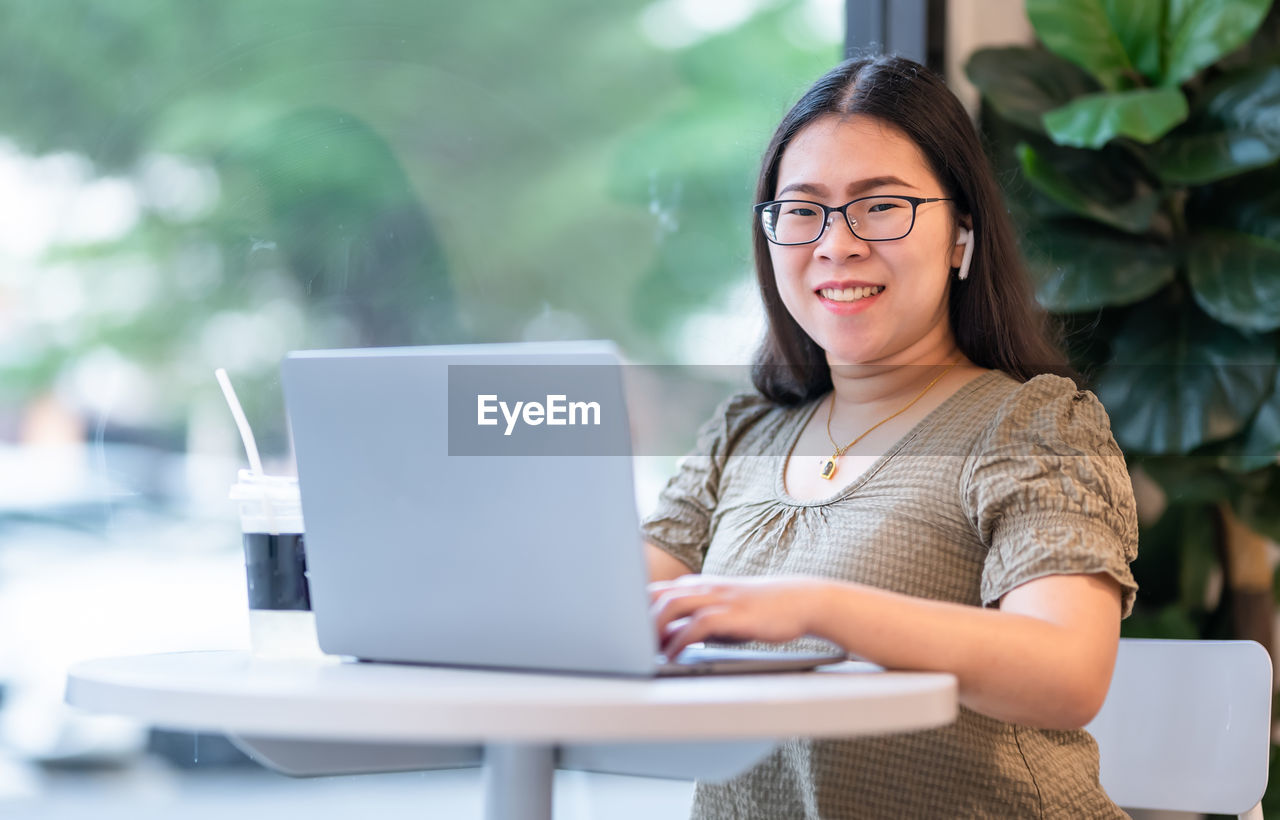 YOUNG WOMAN USING MOBILE PHONE WHILE SITTING ON TABLE AT CAFE