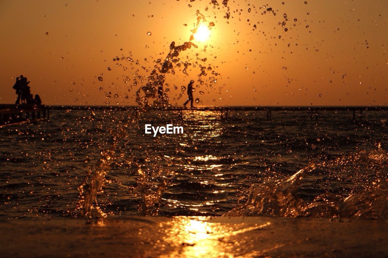 SILHOUETTE OF SPLASHING WATER IN SEA AGAINST SKY DURING SUNSET