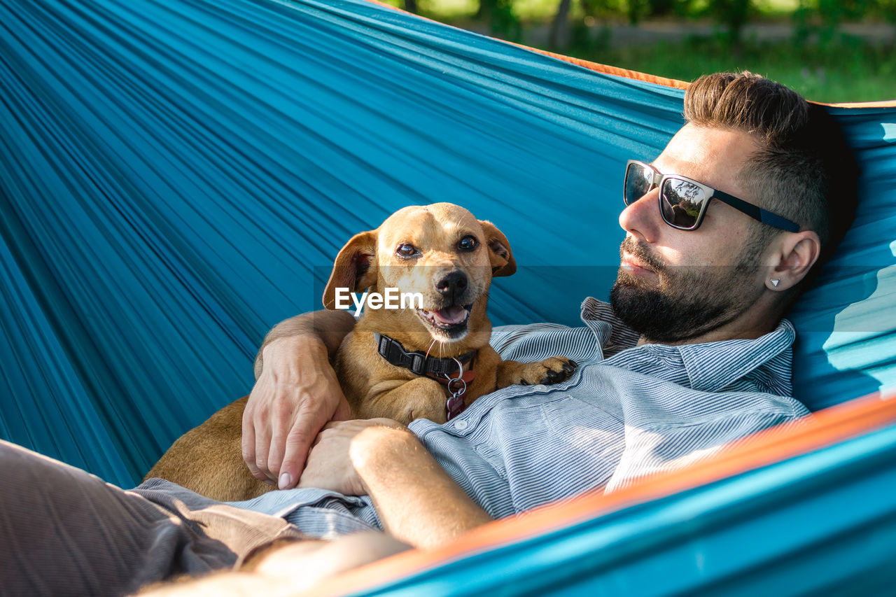 Handsome european man in sunglasses is resting in hammock with his cute little dog.