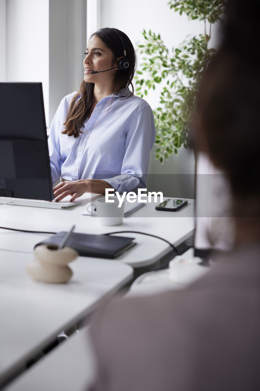 Smiling mid adult businesswoman using headset in office in front of computer screen