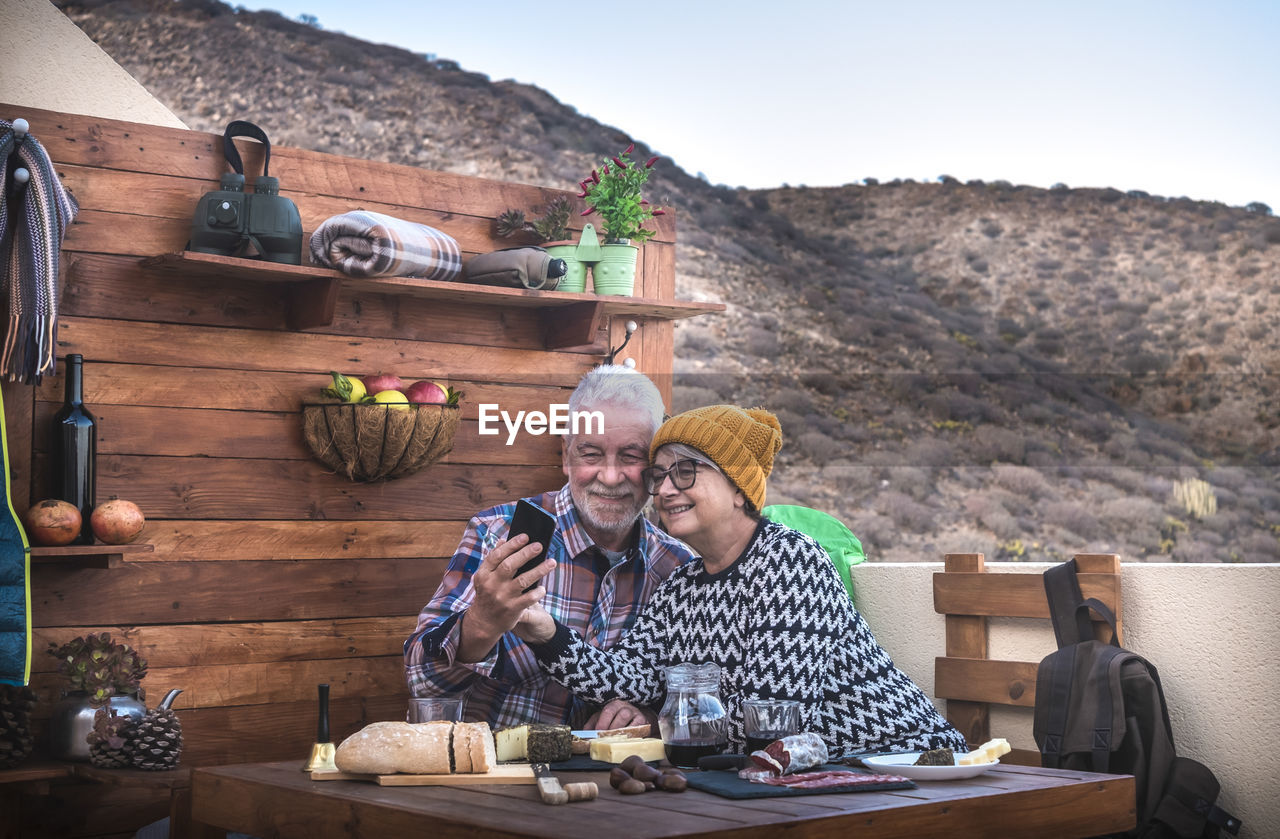 Smiling senior couple sitting at restaurant against mountain and sky