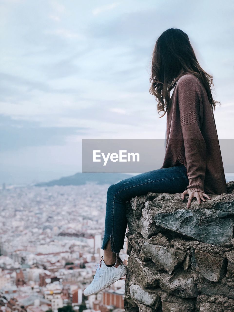 Side view of woman looking at city while sitting on cliff against sky