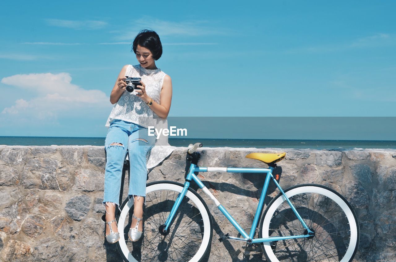 Young woman with bicycle holding camera while sitting on retaining wall at beach against sky