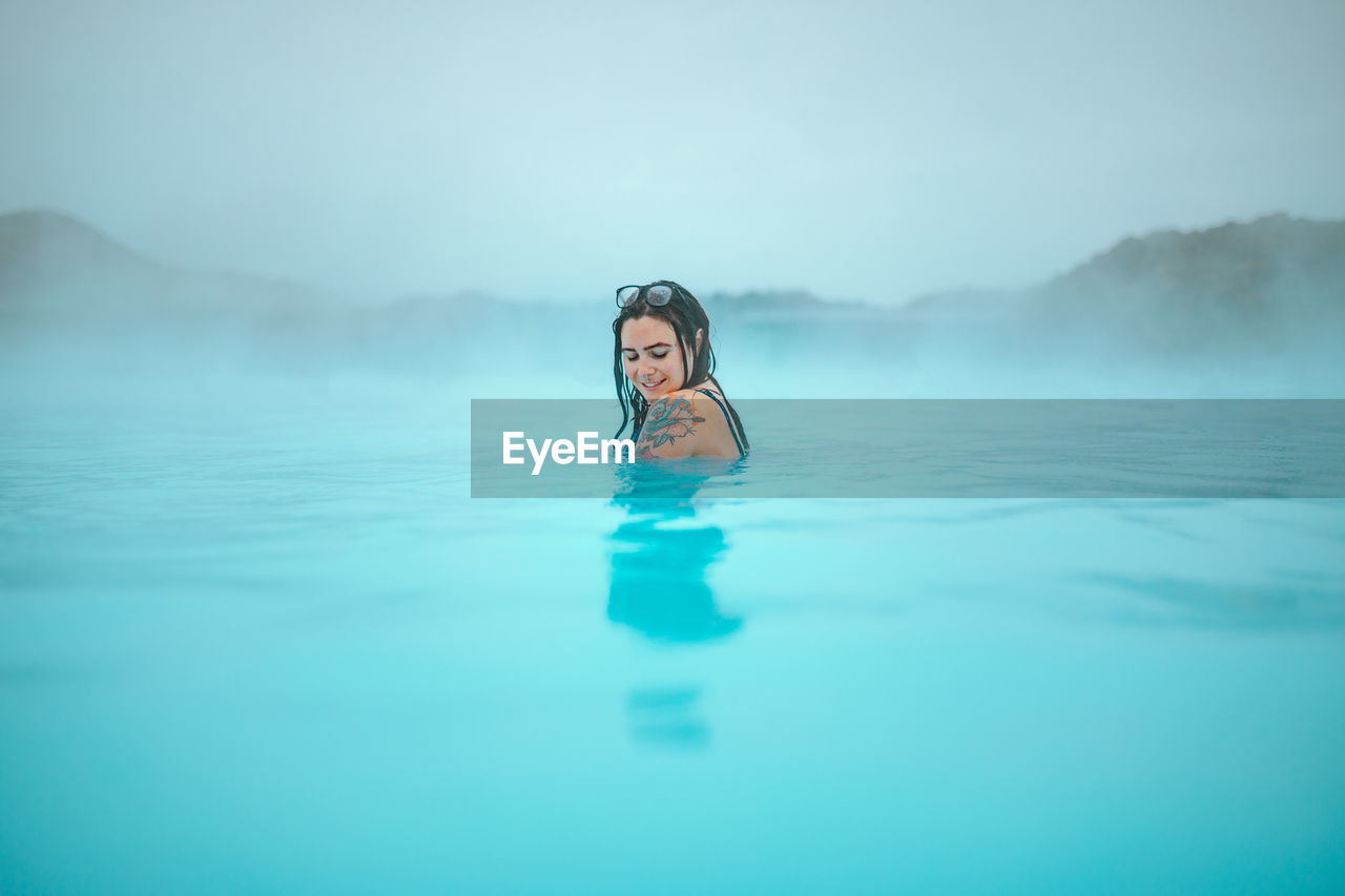 Side view of young tattooed hipster in swimsuit posing in blue water between rocks