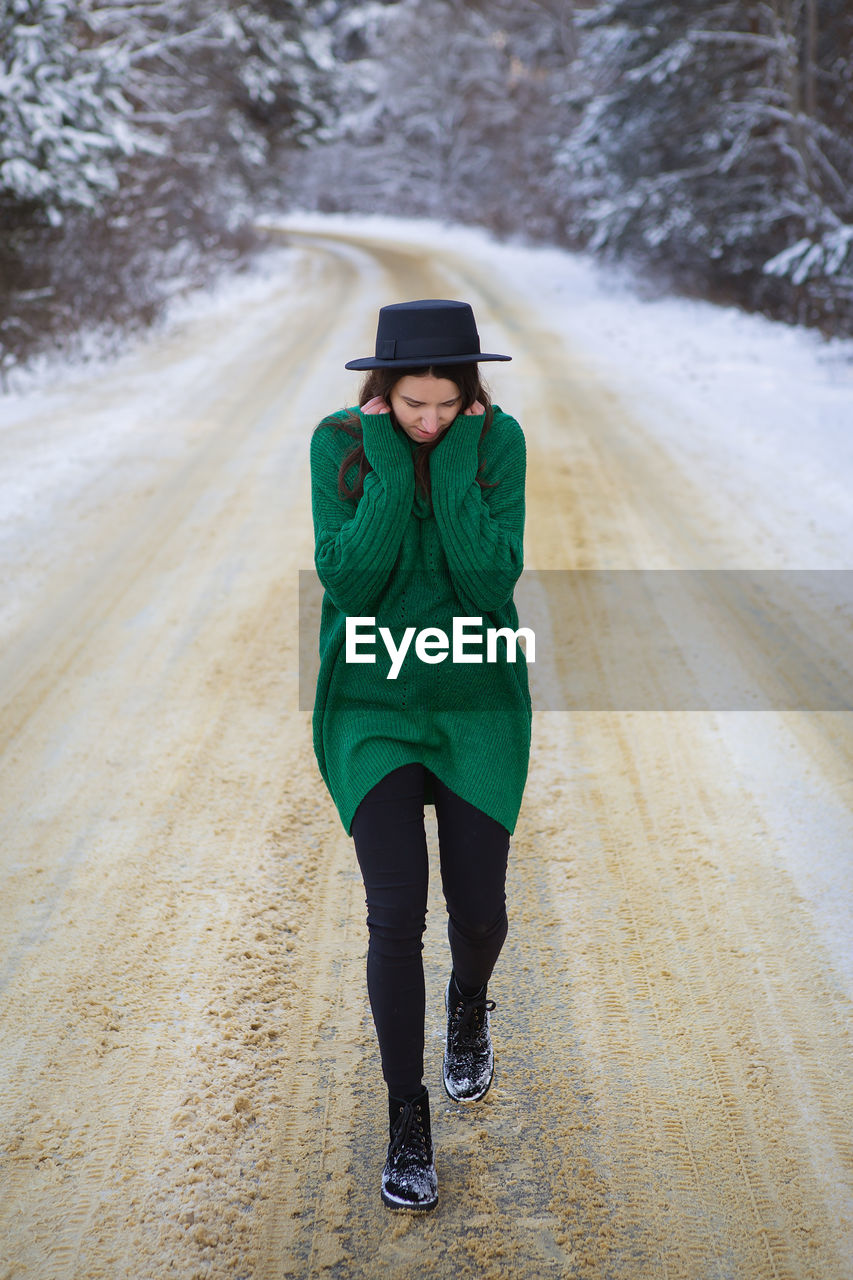 A young girl in a green sweater stands in the middle of a snowy road in a thick pine forest. f