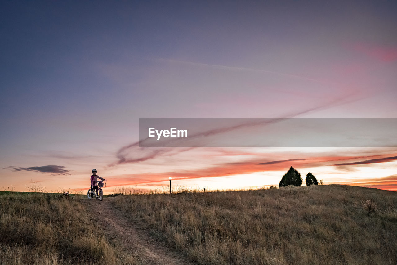 Young girl starts to bike down a dirt path on a grass hill at sunset