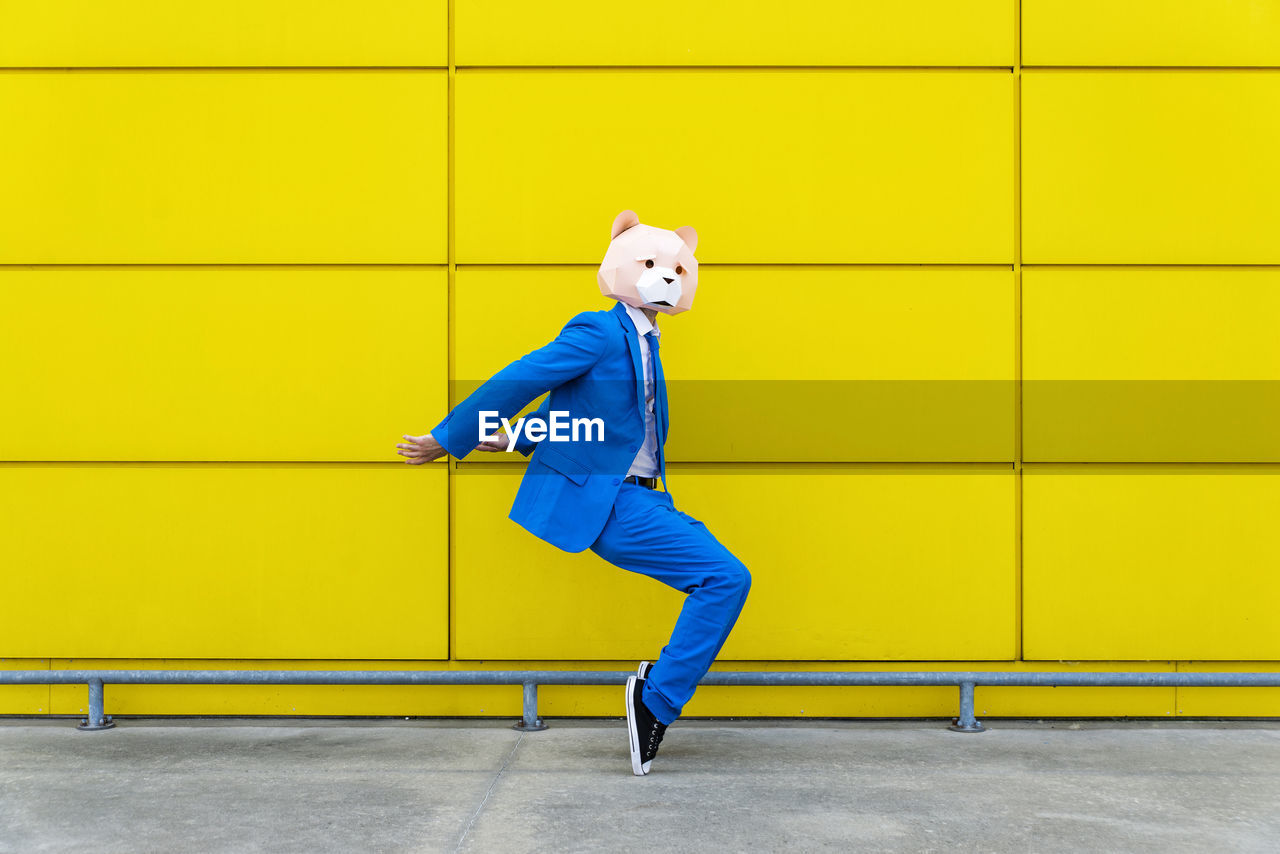 Man wearing vibrant blue suit and bear mask standing en pointe against yellow wall