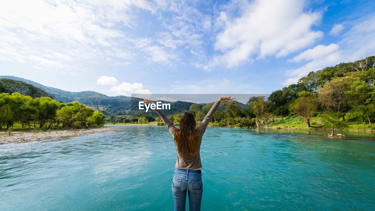 Rear view of woman with arms raised by river against sky