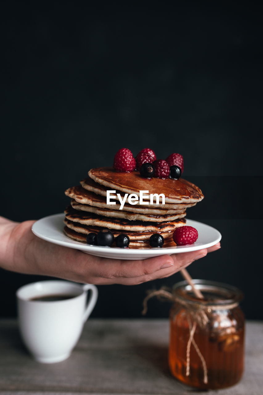 Juicy pancakes with berries and honey on white plate on human hand, jar and spoon, wooden table