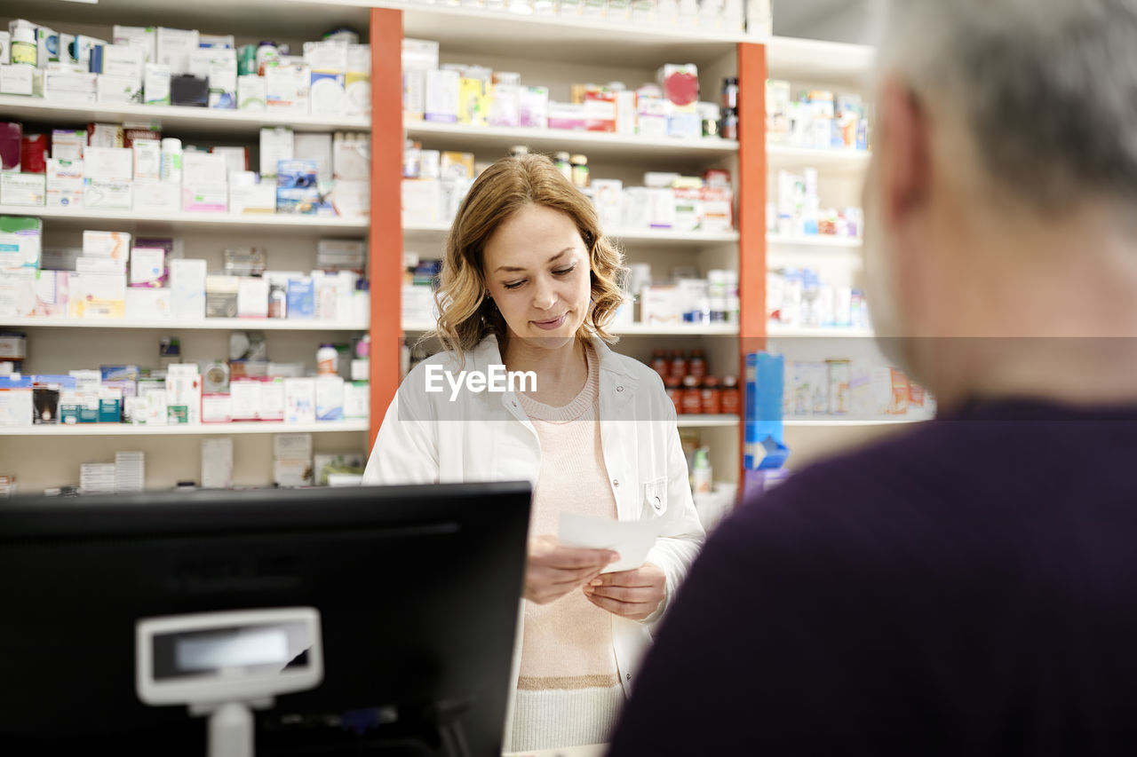 Female pharmacist reading prescription to customer at checkout counter in pharmacy store