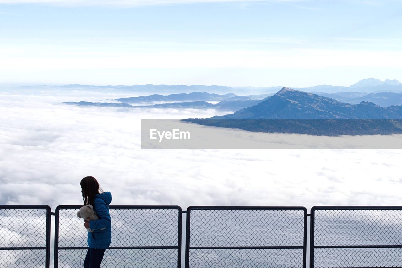 Side view of girl with teddy bear at railing by cloudy sky