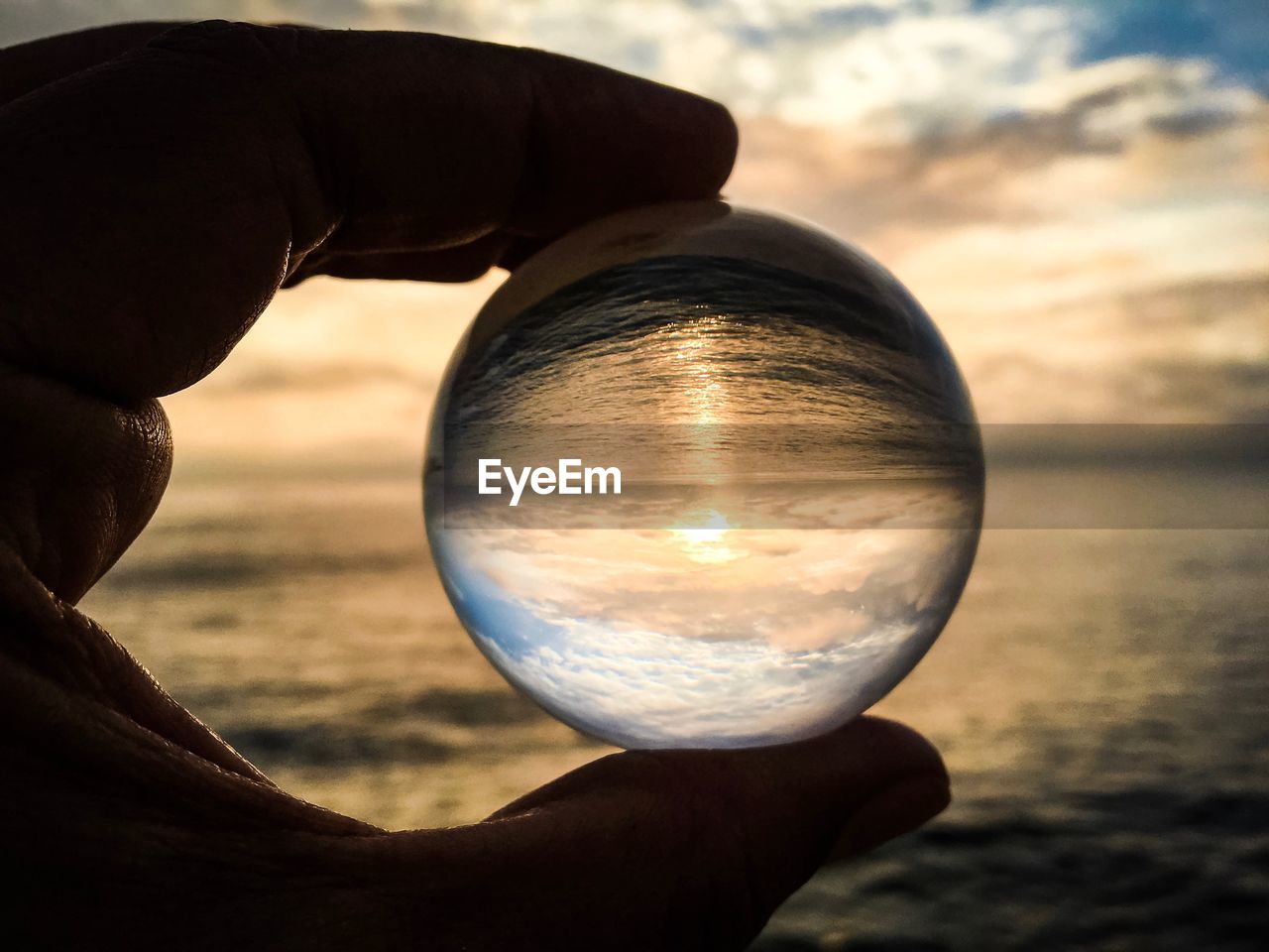 Close-up of hand holding crystal ball against sea at sunset