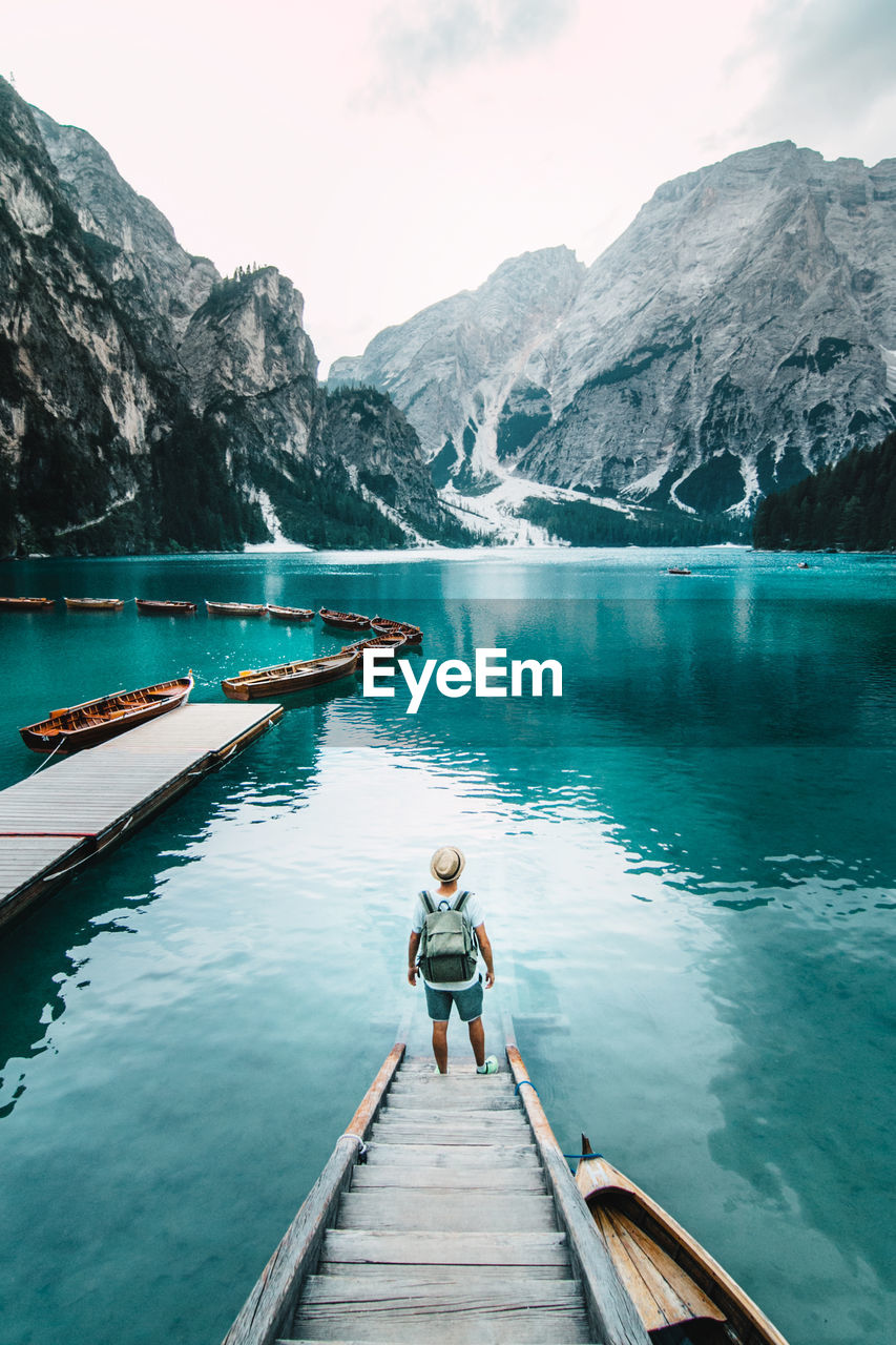 Back view of unrecognizable male traveler standing on wooden quay and admiring amazing scenery of lake with turquoise water in mountains on foggy day