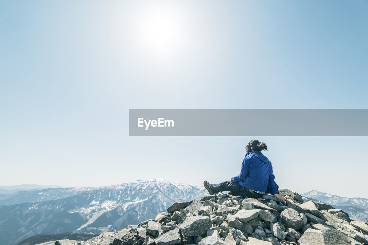 Rear view of man sitting against mountain range against clear sky