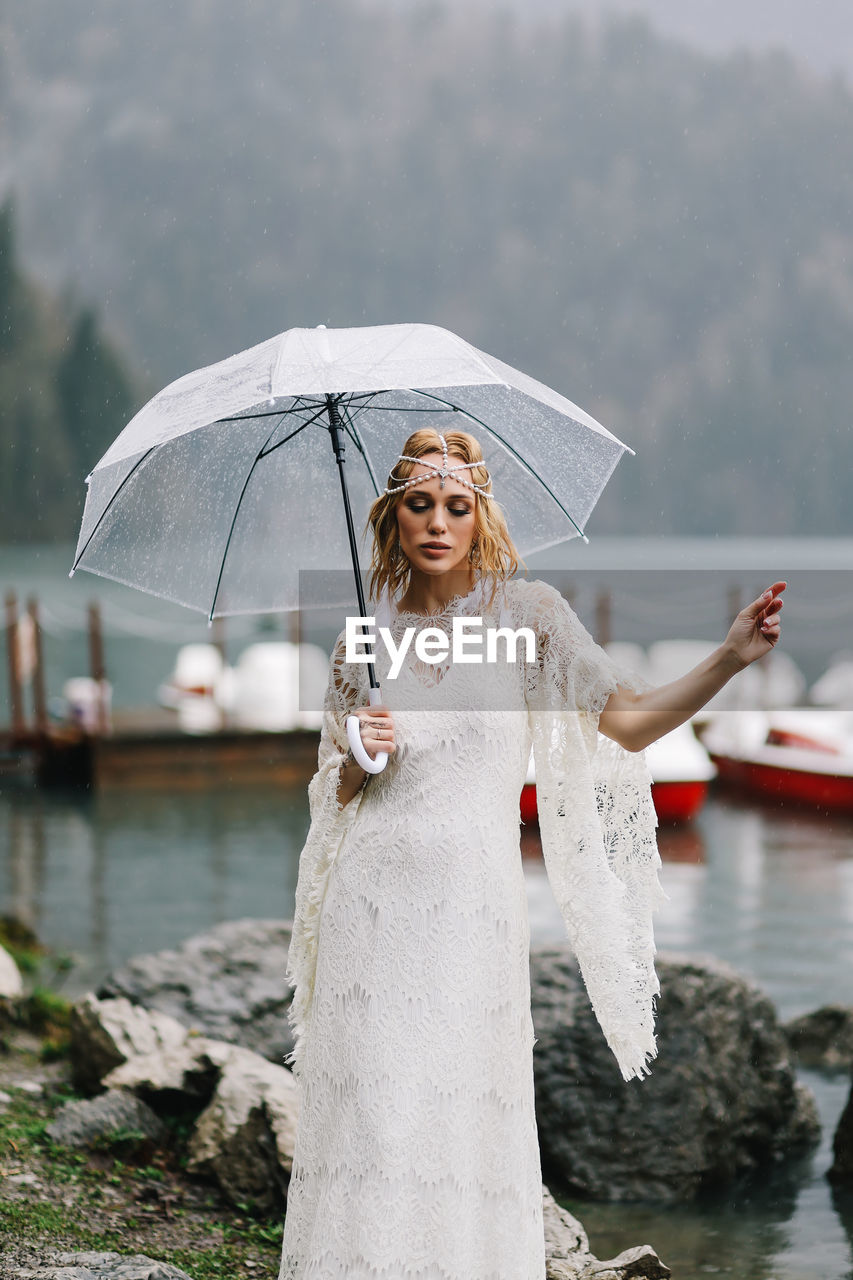 A young woman in a white lace wedding dress stands in the rain among the sea and mountains