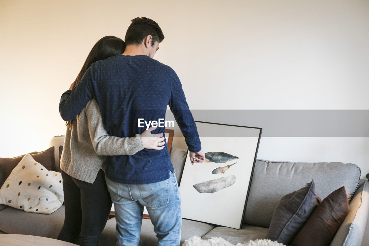 Rear view of young couple embracing while choosing painting on sofa at home