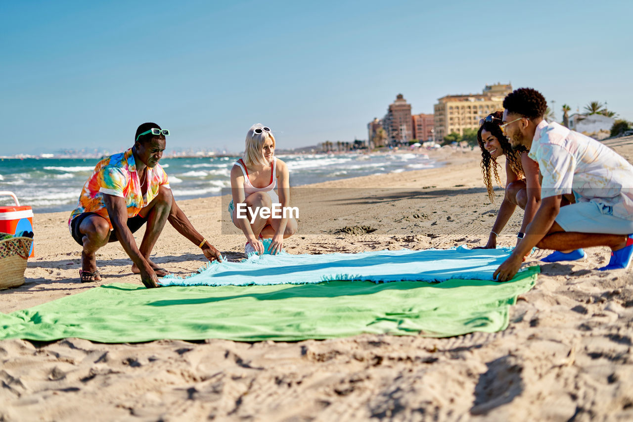 Full body diverse men and women laying and adjusting blanket on sand near sea while spending sunny weekend day on beach in city