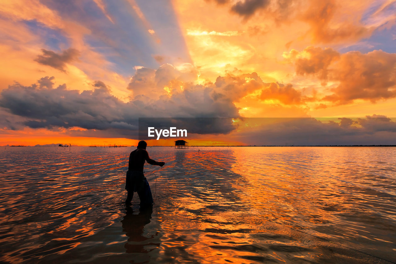 Fisherman fishing in sea against cloudy sky during sunset